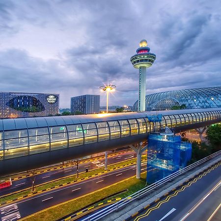 Crowne Plaza Changi Airport By Ihg Singapore Exterior foto The photo depicts an airport scene, likely featuring a modern terminal structure with a distinctive, futuristic design. In the foreground, there is a pedestrian bridge or walkway, along with roads leading to the terminal. A control tower can be seen 