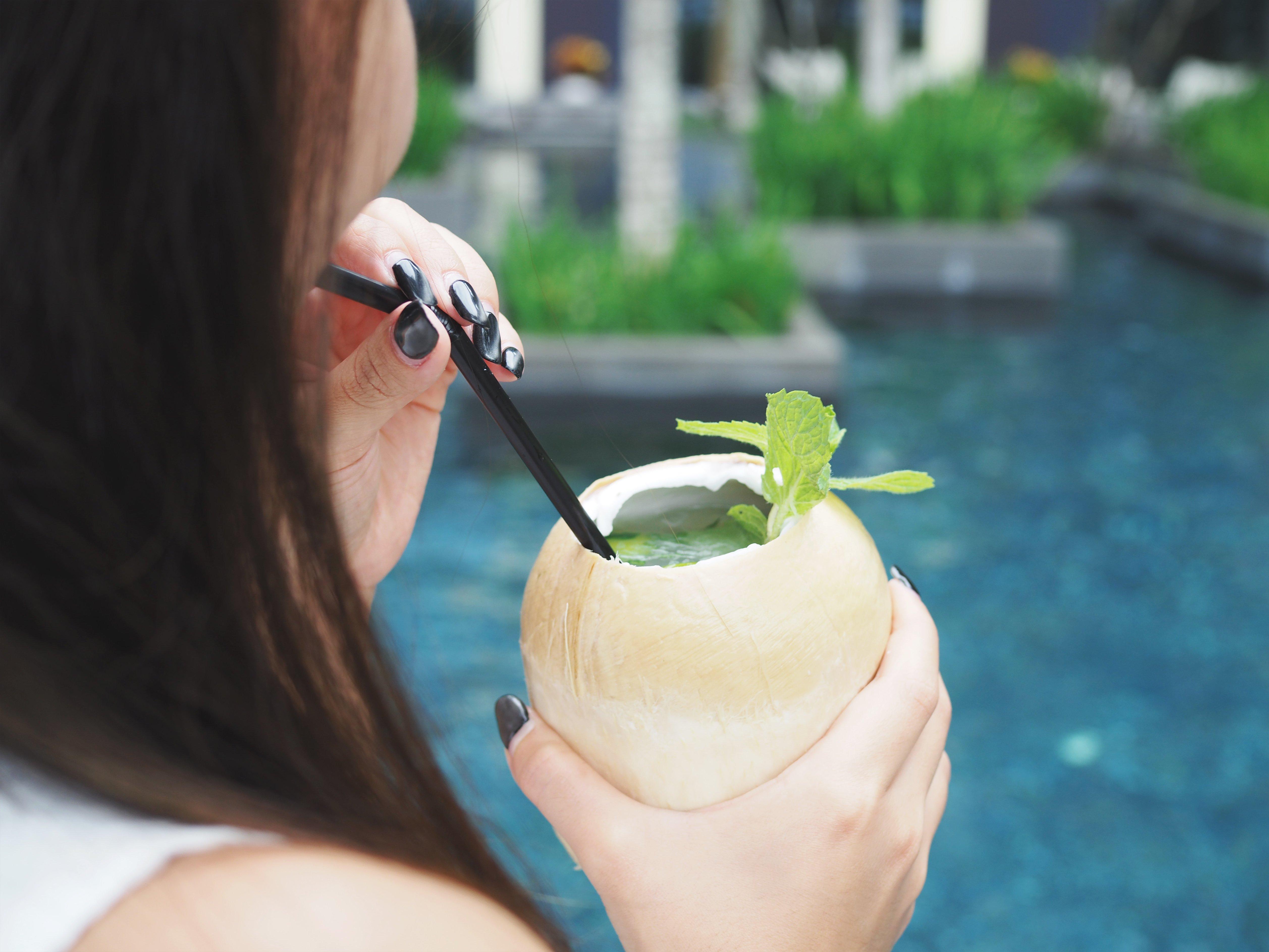 Crowne Plaza Changi Airport By Ihg Singapore Exterior foto The photo features a person holding a coconut drink with a straw. The drink is garnished with a sprig of mint. The background shows a blue pool, suggesting a tropical or relaxing setting. The focus is on the drink and the person's hand.