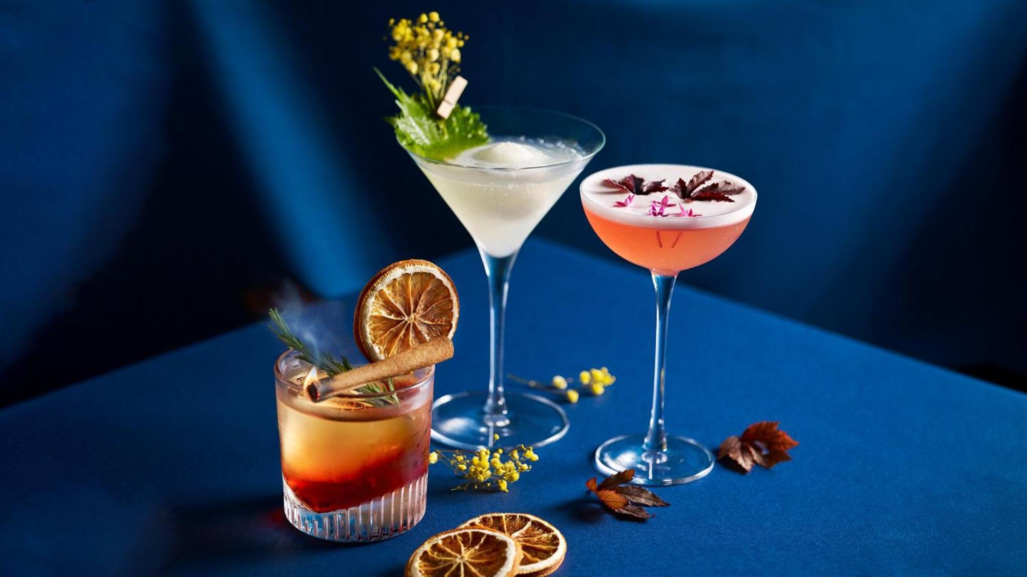 Crowne Plaza Changi Airport By Ihg Singapore Exterior foto The photo shows a stylish arrangement of three cocktails on a blue surface. 

1. **Left Cocktail**: A lowball glass containing a drink with a reddish hue, garnished with a dried orange slice and a cinnamon stick. 

2. **Middle Cocktail**: A classic m