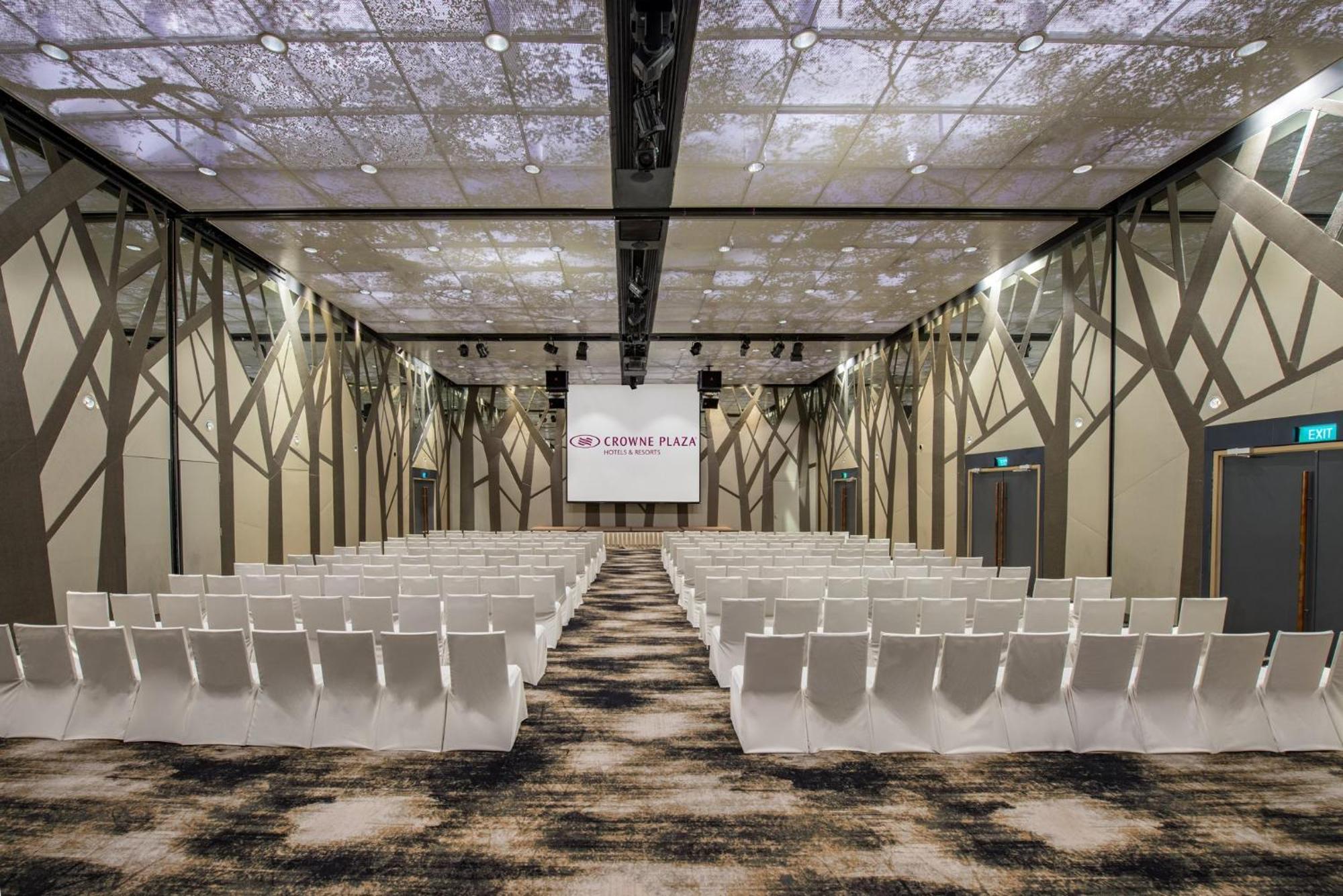 Crowne Plaza Changi Airport By Ihg Singapore Exterior foto The photo shows a modern conference or event space. The room features rows of white chairs arranged in a theater-style layout, facing a large screen with a simple logo. The ceiling is designed to resemble a natural setting, with images that suggest t