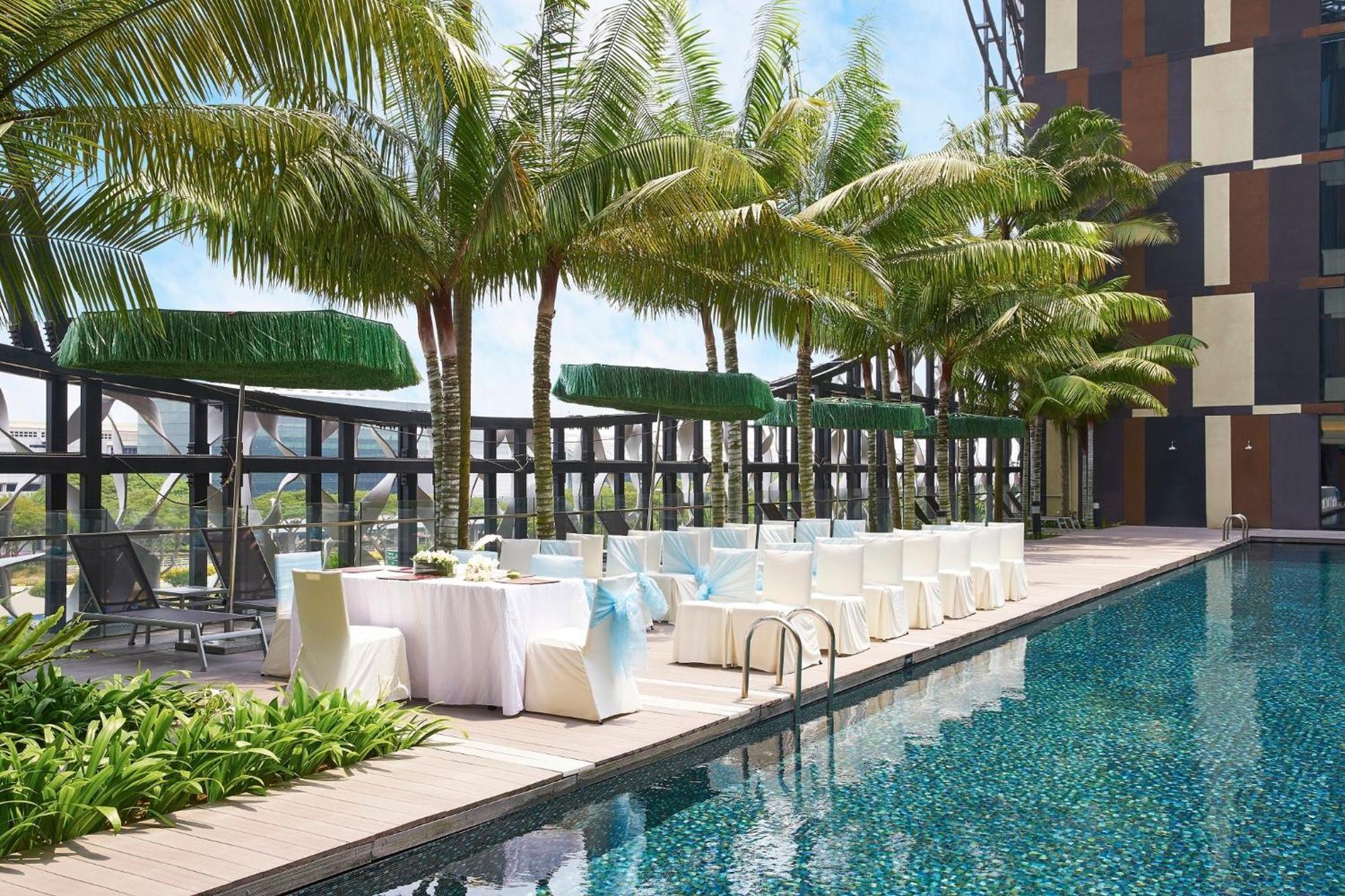 Crowne Plaza Changi Airport By Ihg Singapore Exterior foto The photo shows a swimming pool area with a modern design. There are several palm trees lining the poolside, creating a tropical ambiance. Alongside the pool, there are white chairs and tables set up, possibly for an outdoor dining or event space. Th