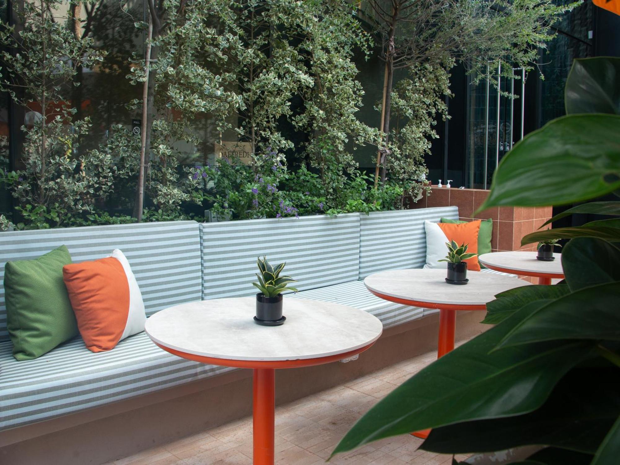 Crowne Plaza Changi Airport By Ihg Singapore Exterior foto The photo shows a modern outdoor seating area. There is a long bench with a corrugated metal backrest, decorated with colorful cushions in green and orange. In front of the bench, there are circular tables with white marble tops and orange legs. Each