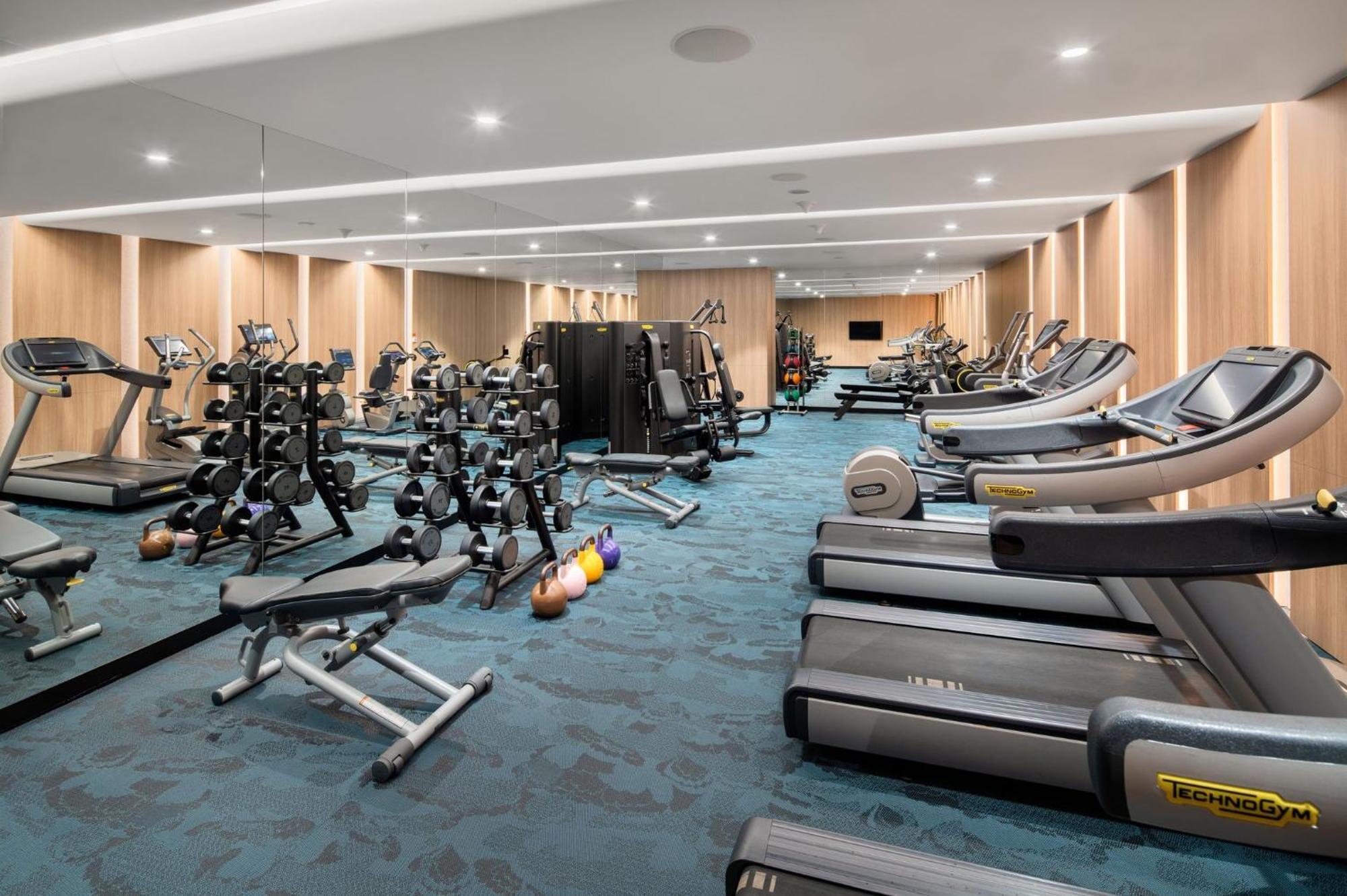 Crowne Plaza Changi Airport By Ihg Singapore Exterior foto The photo shows a modern gym interior. It features various fitness equipment, including multiple treadmills, weightlifting machines, and a set of free weights on racks. The flooring has a blue carpet, and the walls are made of wood paneling, giving t